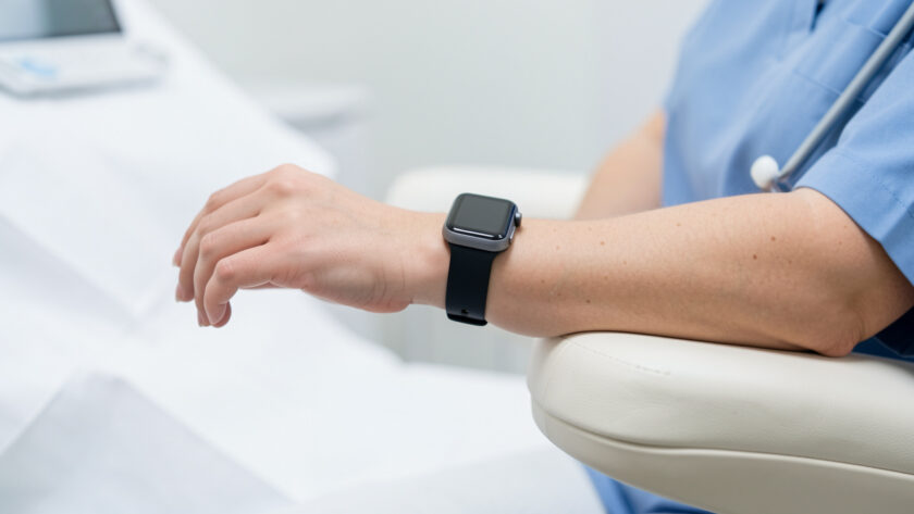 Gros plan réaliste d’une montre connectée portée au poignet d’une femme assise, le bras posé à hauteur du cœur dans un environnement clinique sobre.